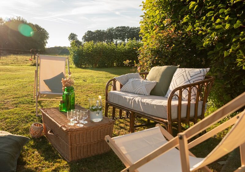 Location d'un salon avec canapé en rotin pour événements, mariage, anniversaires, soirée d'été dans un thème bohème chic et champêtre. Location de chilienne beige, malle en osier vintage et coussins décoratifs